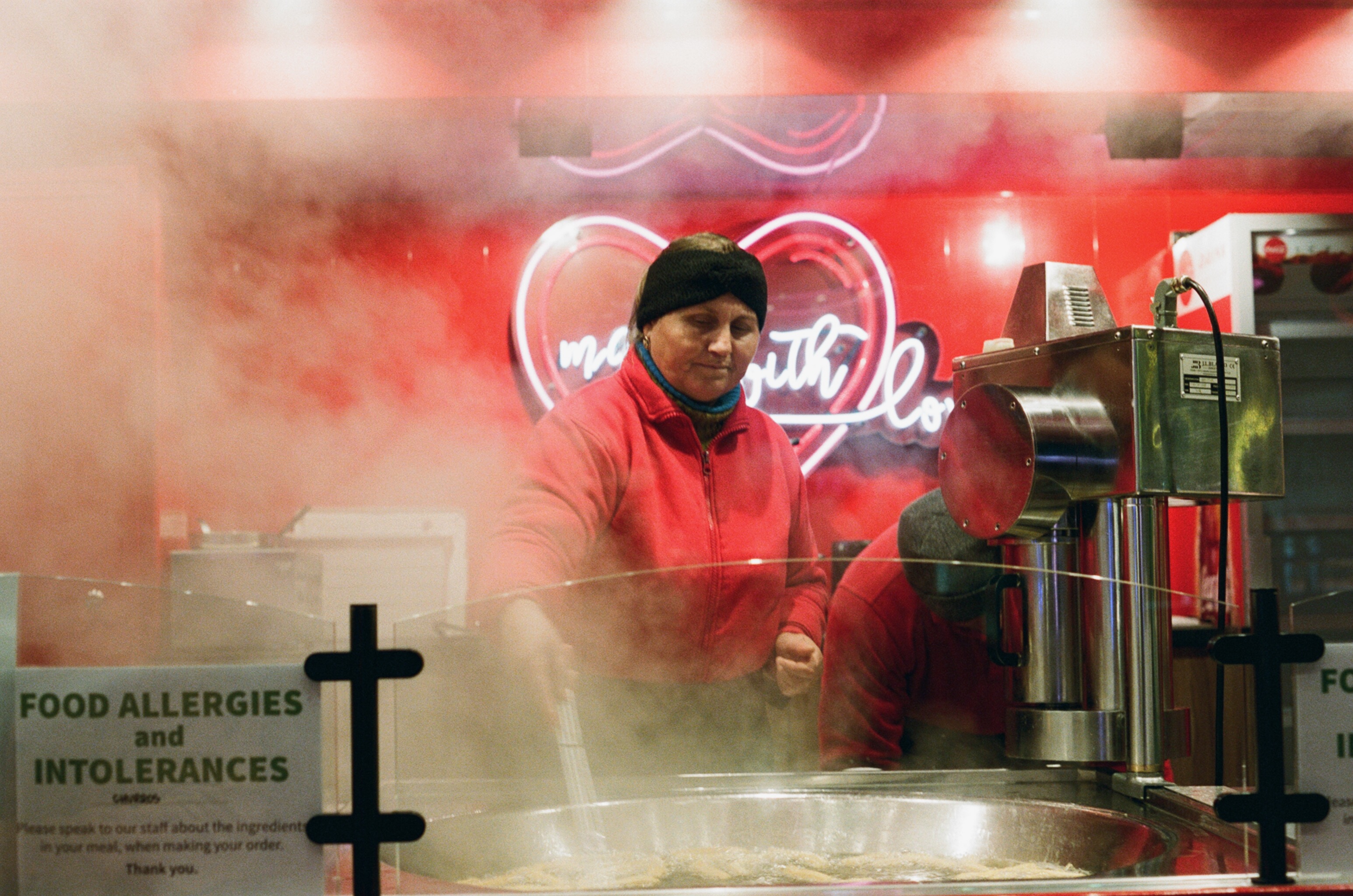 A woman working at a food counter, seen through glass clouded with steam, a neon sign glowing behind her.