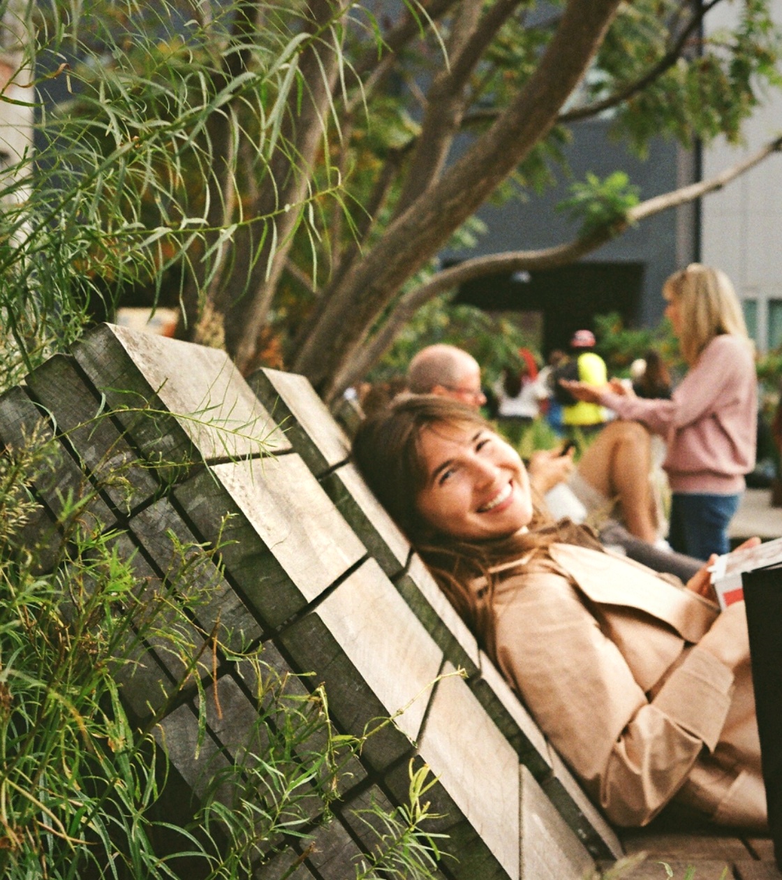 A woman reclining on a wooden bench under planting, smiling toward the camera.