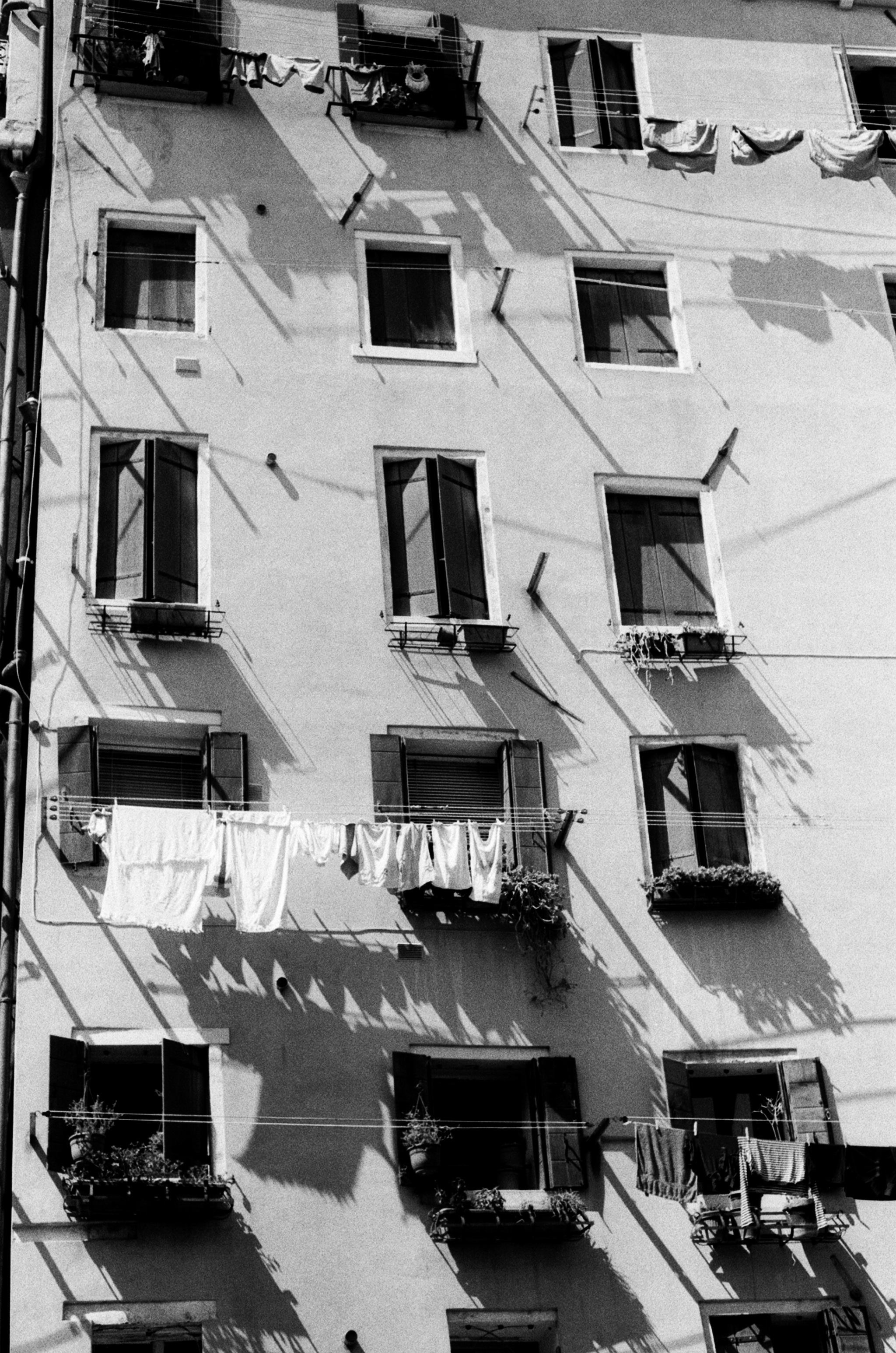 A tall pale facade of shuttered windows, crossed by hard diagonal shadows and lines of drying laundry.