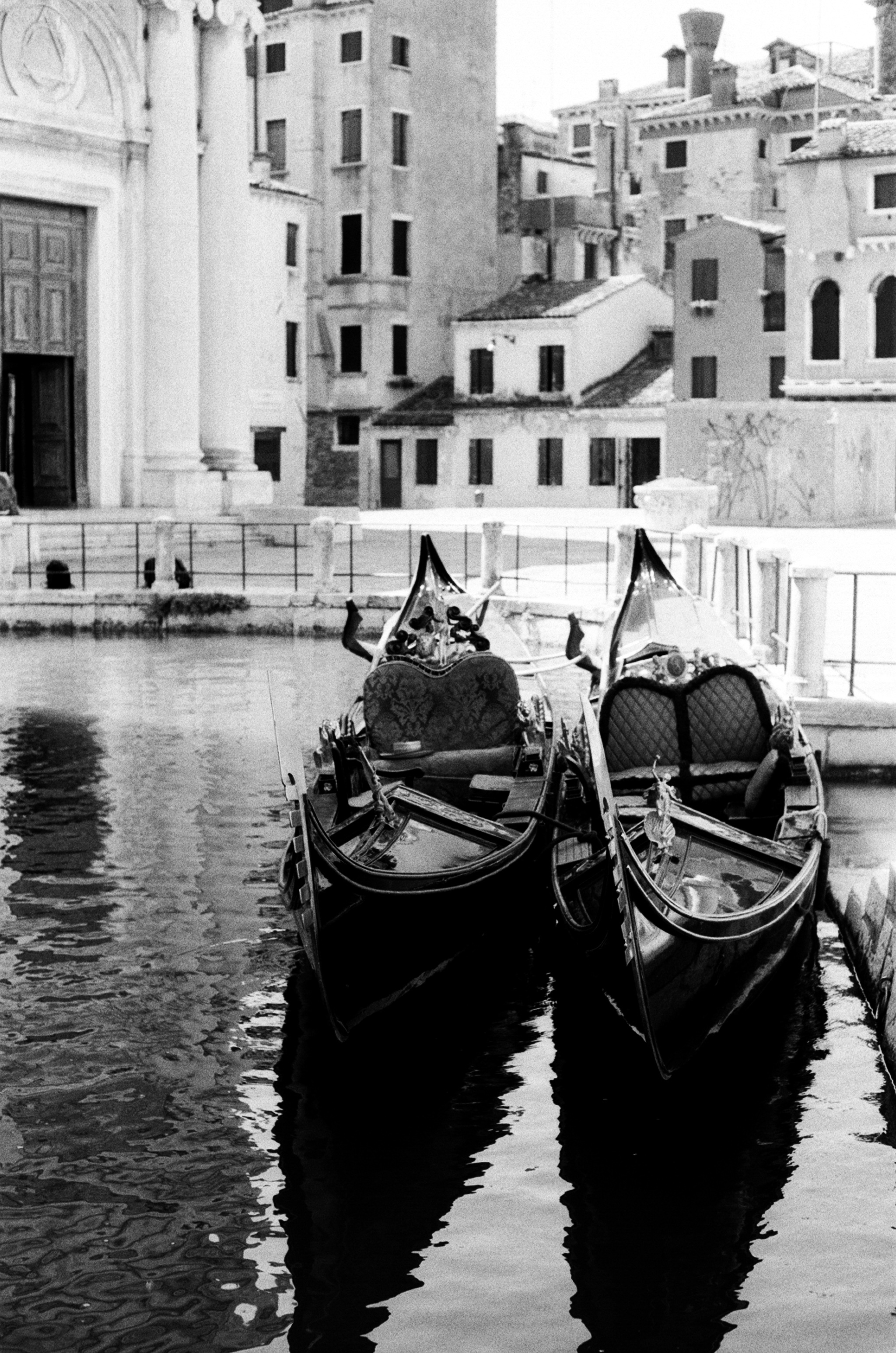 Two moored gondolas seen bow-on beside a church facade, their forms doubled in the water.