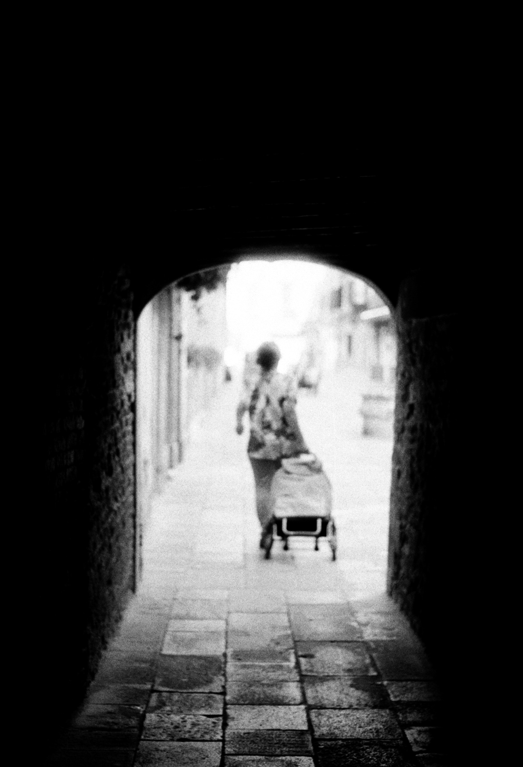 A dark archway opening onto bright daylight, a woman with a shopping trolley walking out into the glare.