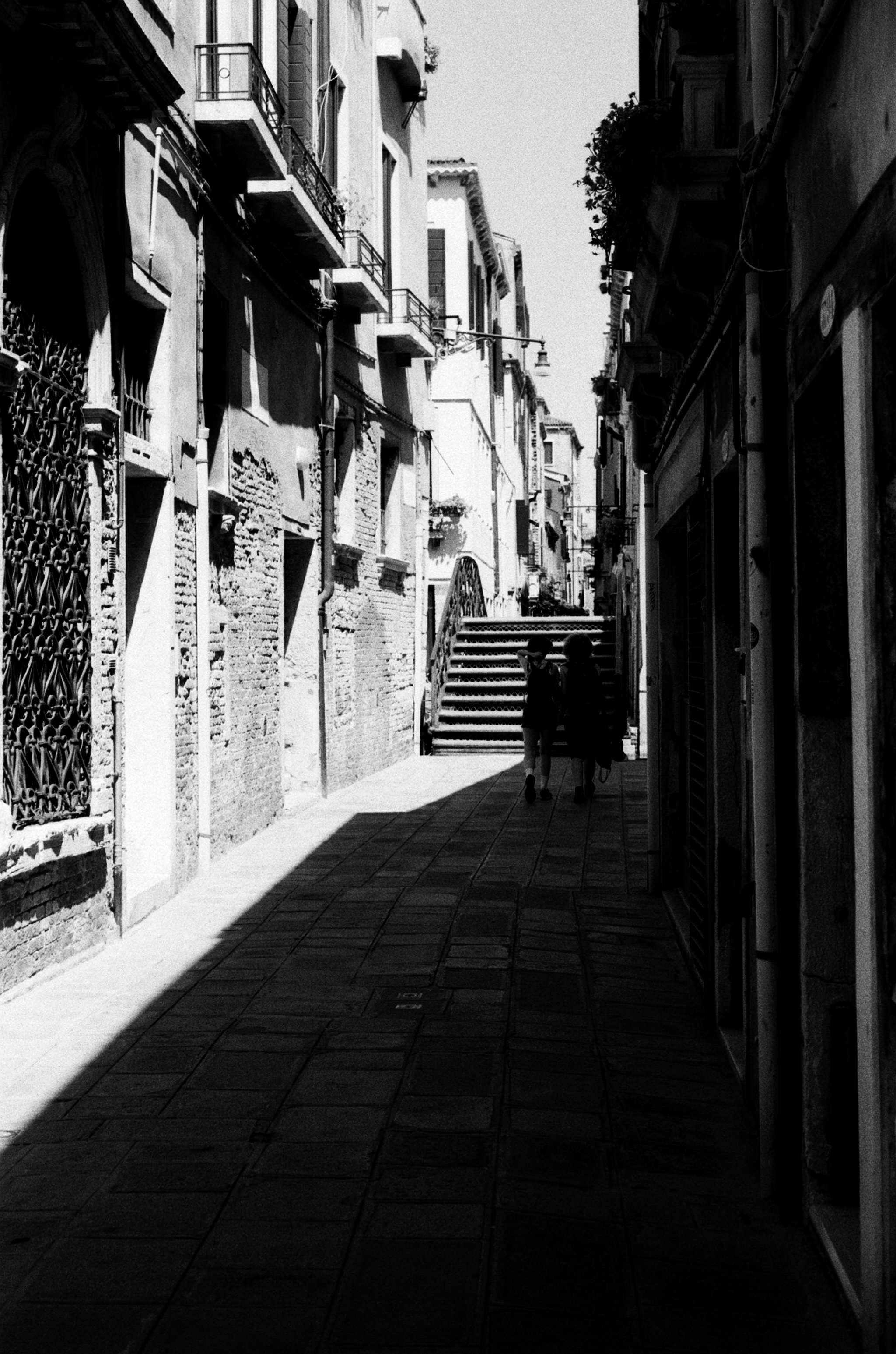 Two figures silhouetted at the far end of a narrow alley, a hard wedge of sunlight cutting the paving.