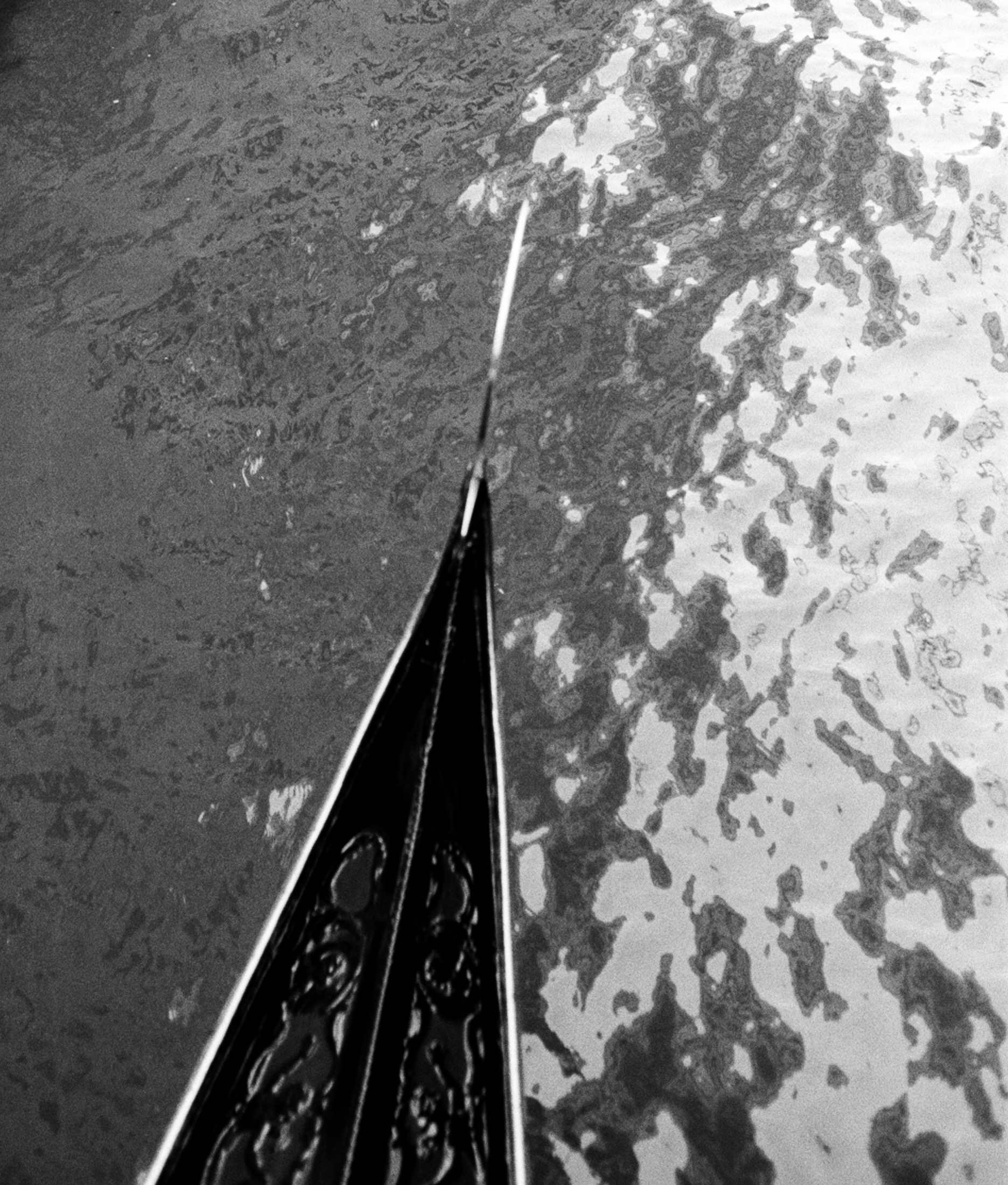The carved black prow of a gondola cutting a pale, mottled expanse of water.