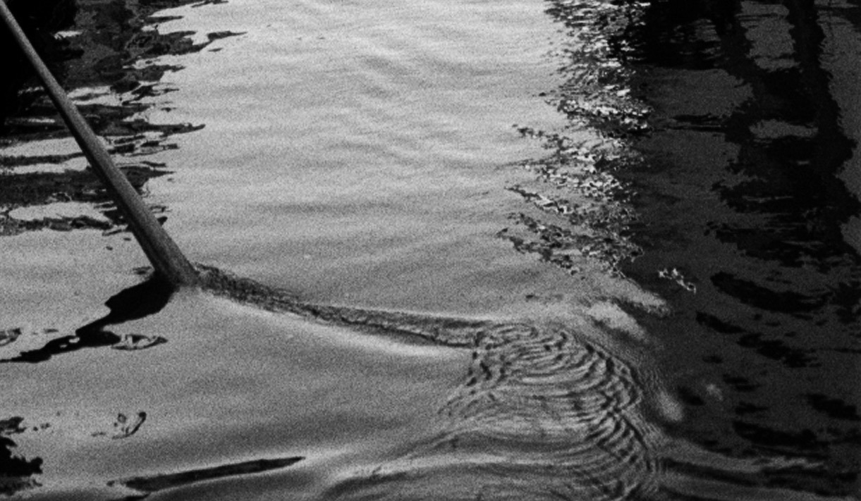 The oar entering the water, drawing a turning vortex of ripples across a pale surface.