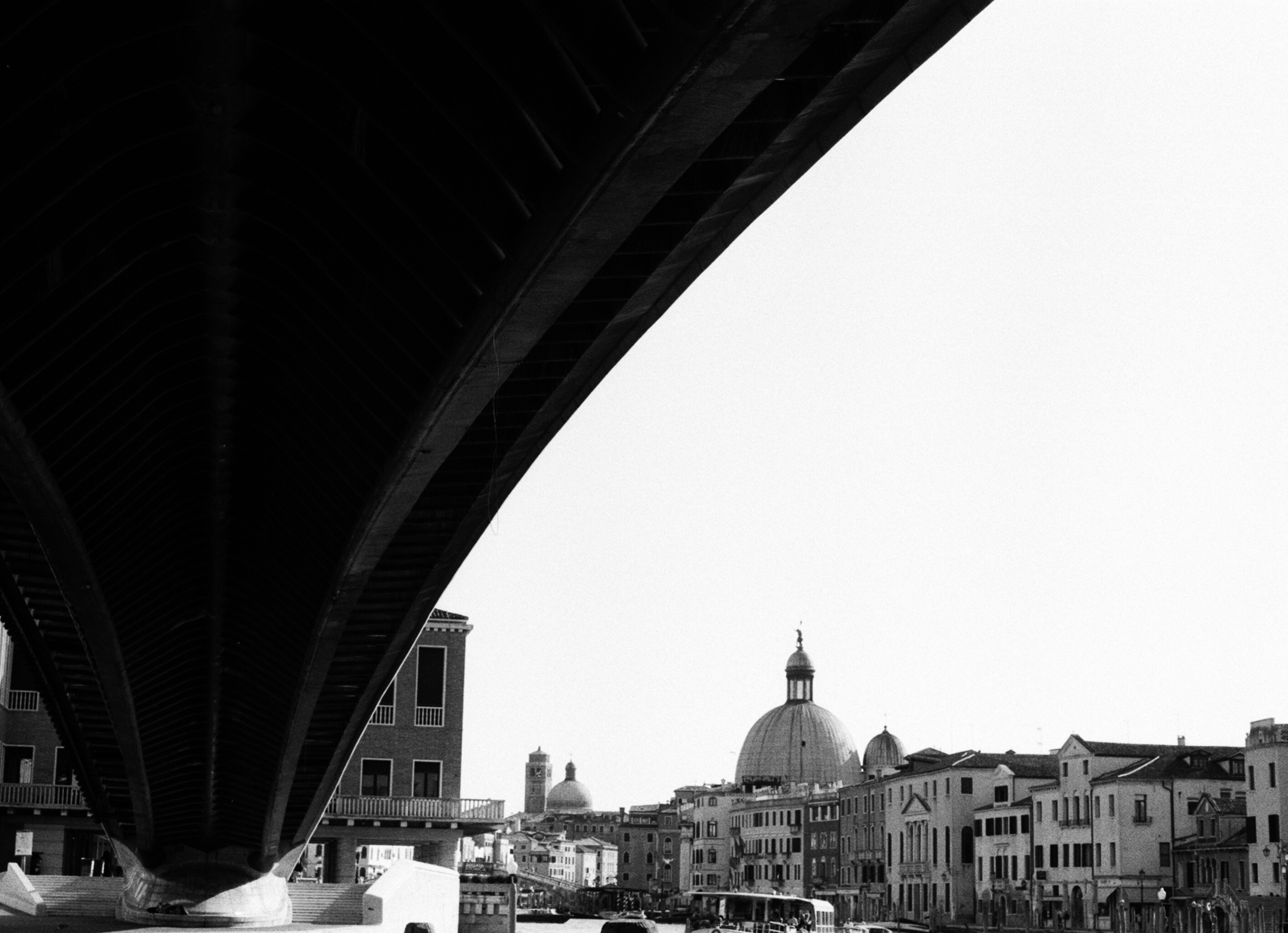 The ribbed underside of the Costituzione bridge sweeping across a white sky, with a dome on the canal below.